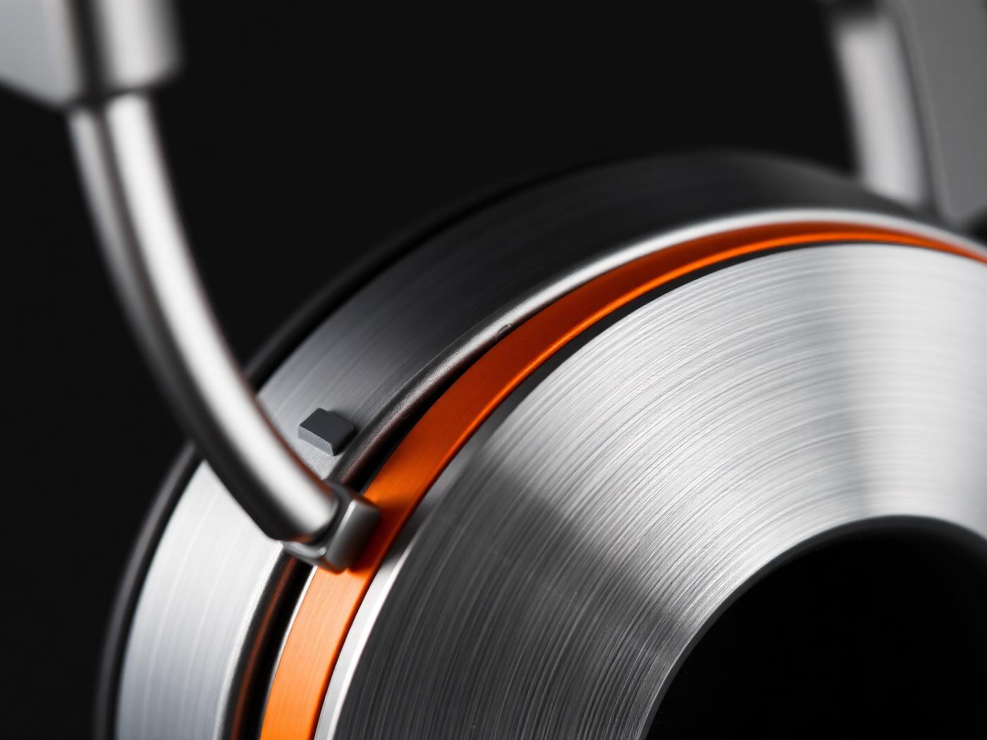 Close detail of an AKOUE headphone earcup: brushed pearl-silver aluminium meeting a single AKOUE Orange accent ring.